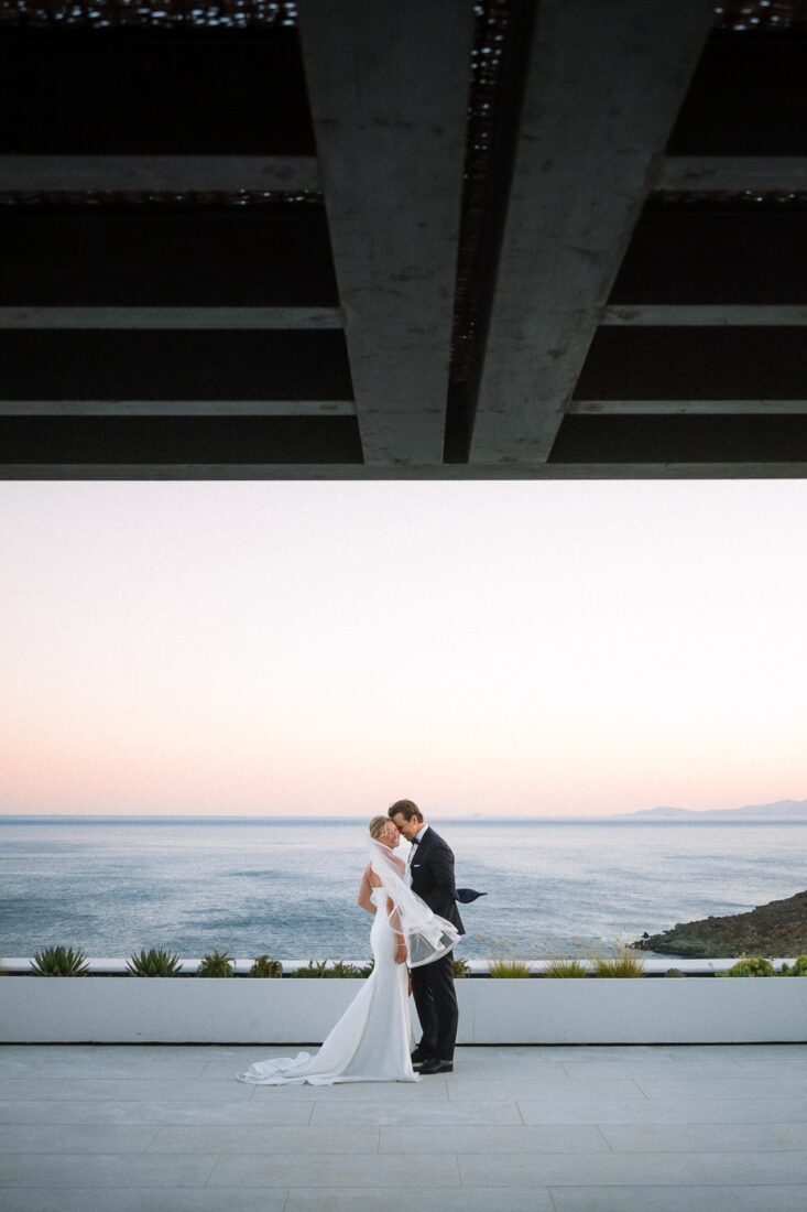 Haley and Mitch embracing on a terrace above the sea at dusk, framed by a dark concrete canopy