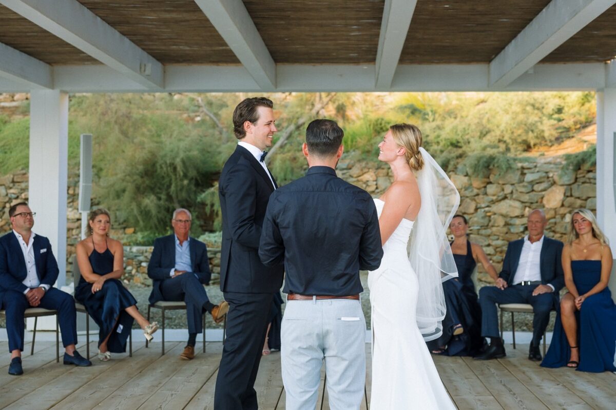 Mitch and Haley smile at each other during their ceremony while their family watches from chairs against a stone wall