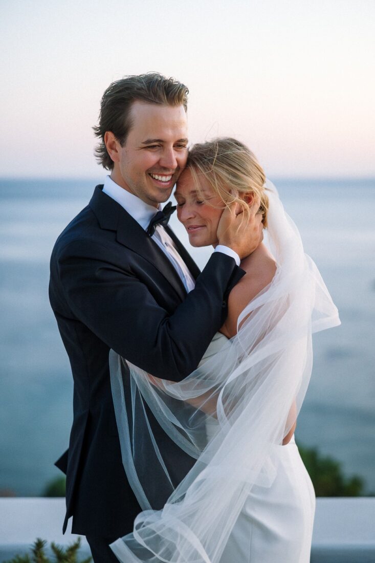 Mitch holds Haley close on a terrace above the sea at dusk, her veil lifting in the wind